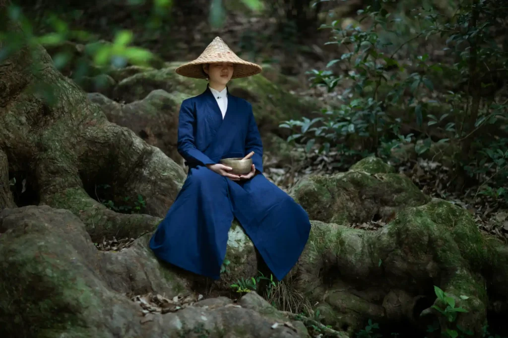 modern asian woman cdressing dark blue kimono holding tibetan bowls in her hands sitting on tree roots with asian hat